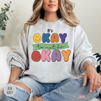 Ash grey sweatshirt with a retro mental health design featuring smiley faces and colorful lettering that says "It's Okay to Not Be Okay."