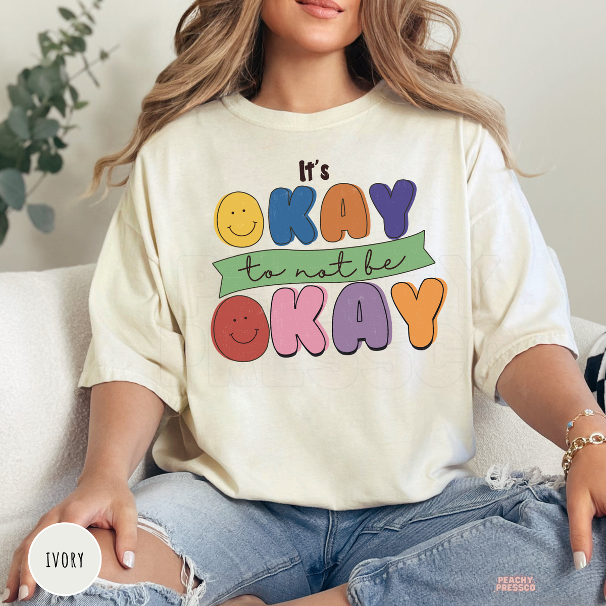 Ivory Comfort Colors t-shirt with a colorful retro smiley face graphic and bold typography reading "It's Okay to Not Be Okay," celebrating kindness and mental wellness.