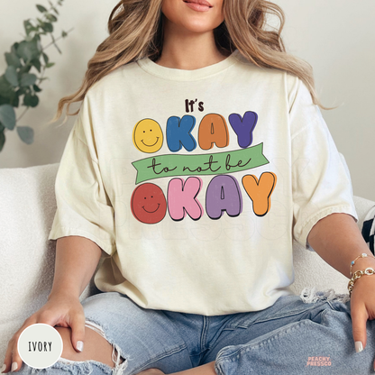 Ivory Comfort Colors t-shirt with a colorful retro smiley face graphic and bold typography reading "It's Okay to Not Be Okay," celebrating kindness and mental wellness.