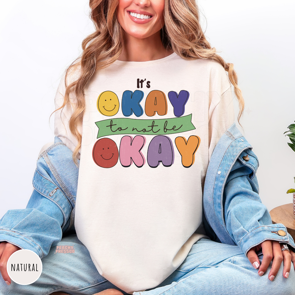 Natural shirt featuring a cheerful retro design with smiley faces and colorful bubble letters displaying the uplifting message "It's Okay to Not Be Okay."