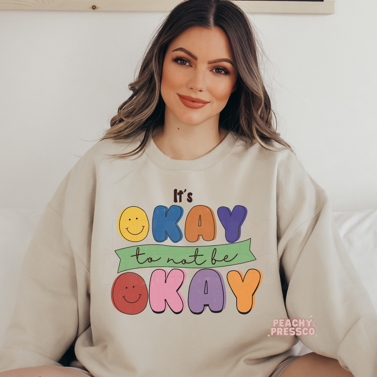 Sand crewneck sweatshirt with a colorful vintage-inspired "It's Okay to Not Be Okay" graphic featuring smiley faces and playful retro typography.