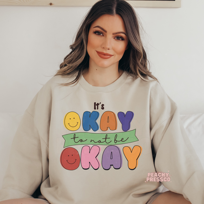 Sand crewneck sweatshirt with a colorful vintage-inspired "It's Okay to Not Be Okay" graphic featuring smiley faces and playful retro typography.