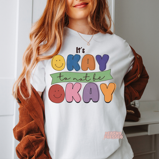 White t-shirt featuring colorful retro bubble lettering with smiley faces that reads "It's Okay to Not Be Okay," promoting mental health awareness and self-compassion.