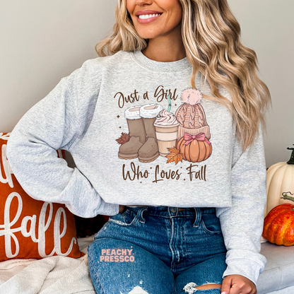Woman wearing an ash grey crewneck sweatshirt with a "Just a Girl Who Loves Fall" design featuring cozy boots, iced pumpkin coffee, knit beanie, pumpkin, and fall aesthetic artwork.