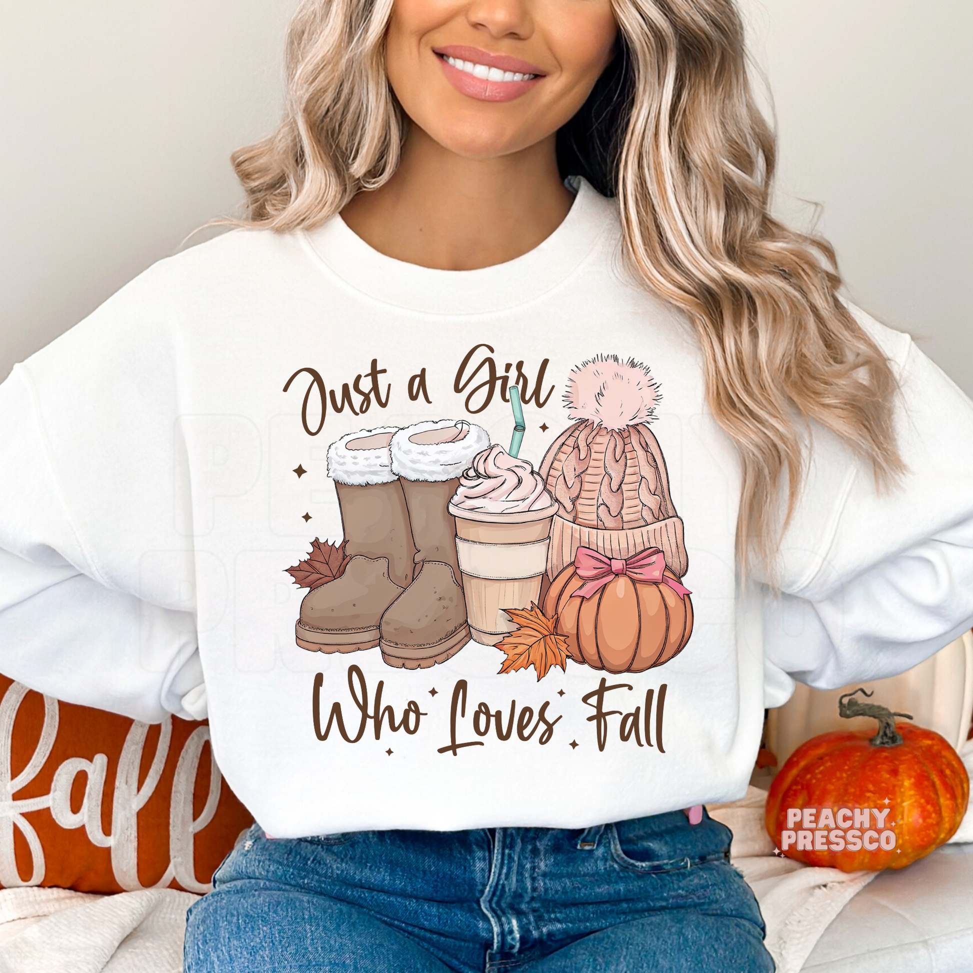 Woman wearing a white crewneck sweatshirt featuring a "Just a Girl Who Loves Fall" graphic with cozy boots, a pumpkin spice coffee, knit beanie, pumpkin, and autumn leaves.