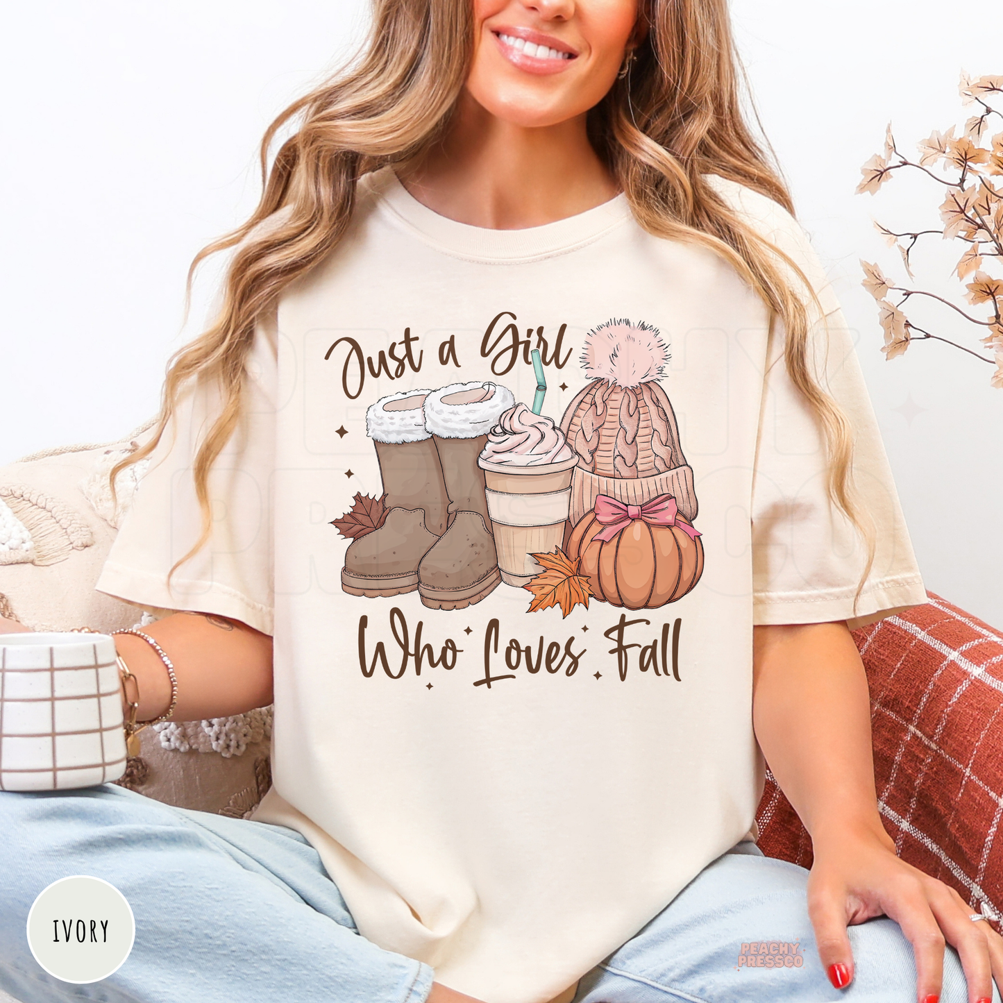 Woman wearing an ivory graphic t-shirt featuring a "Just a Girl Who Loves Fall" design with cozy boots, pumpkin spice coffee, pink knit hat, pumpkin, and autumn leaves.