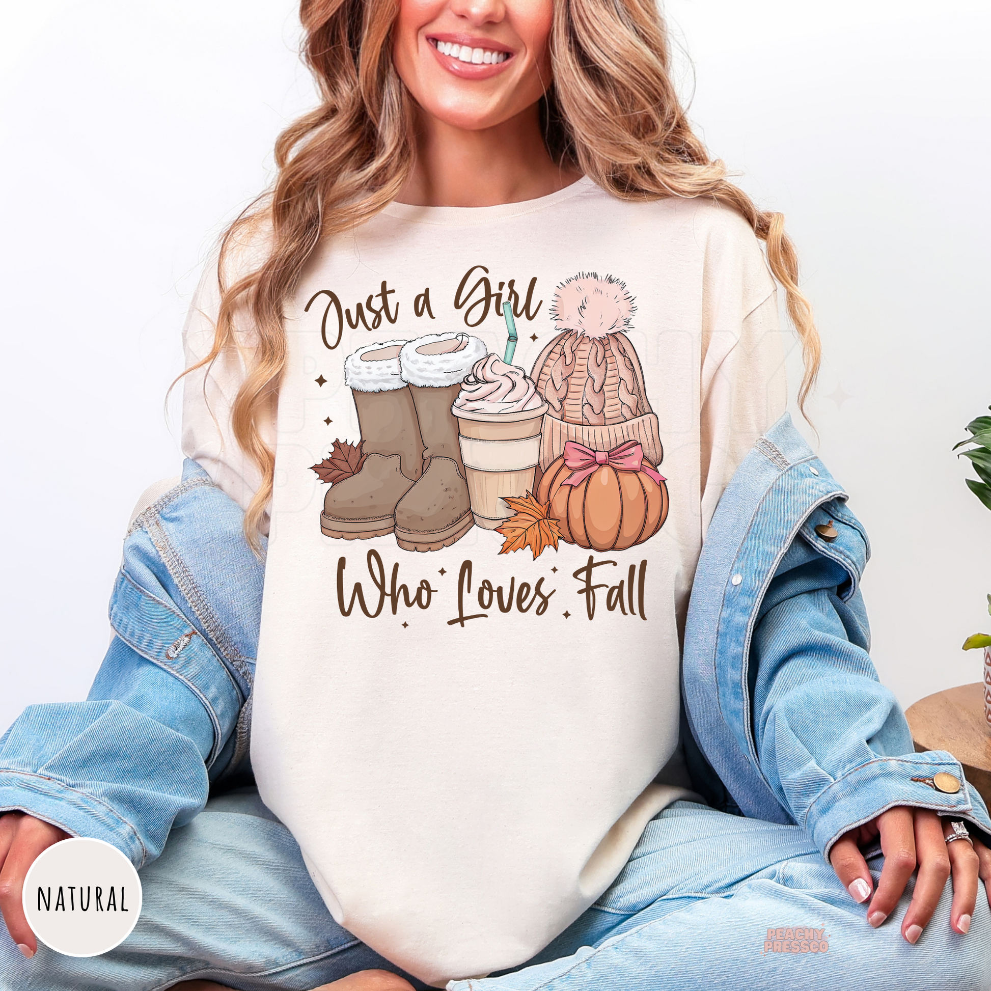 Woman wearing a natural graphic t-shirt featuring a "Just a Girl Who Loves Fall" design with cozy boots, iced coffee, knit beanie, pumpkin, and cute fall aesthetic illustration.