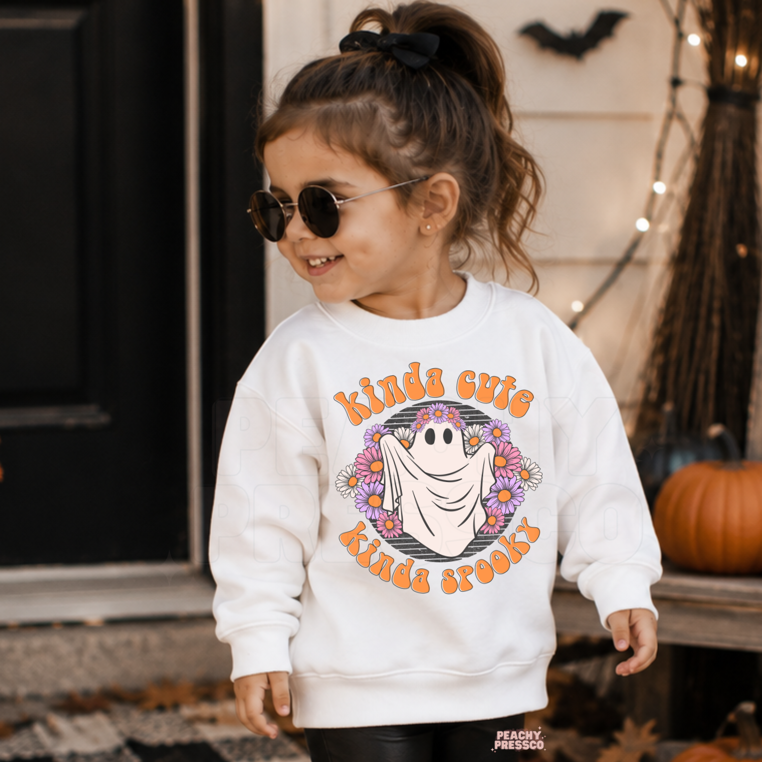Girl wearing a white Kinda Cute Kinda Spooky Halloween sweatshirt featuring a cute floral ghost graphic in a fall porch setting.