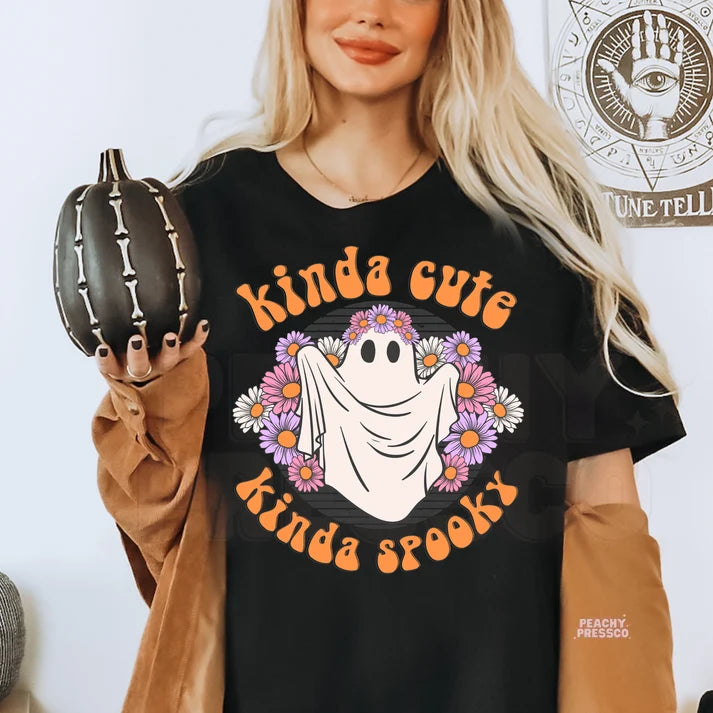 A woman wearing a black graphic t-shirt with a 'kinda cute kinda spooky' ghost and flower design, holding a small black pumpkin. The t-shirt features orange text and a white ghost surrounded by purple and pink flowers.