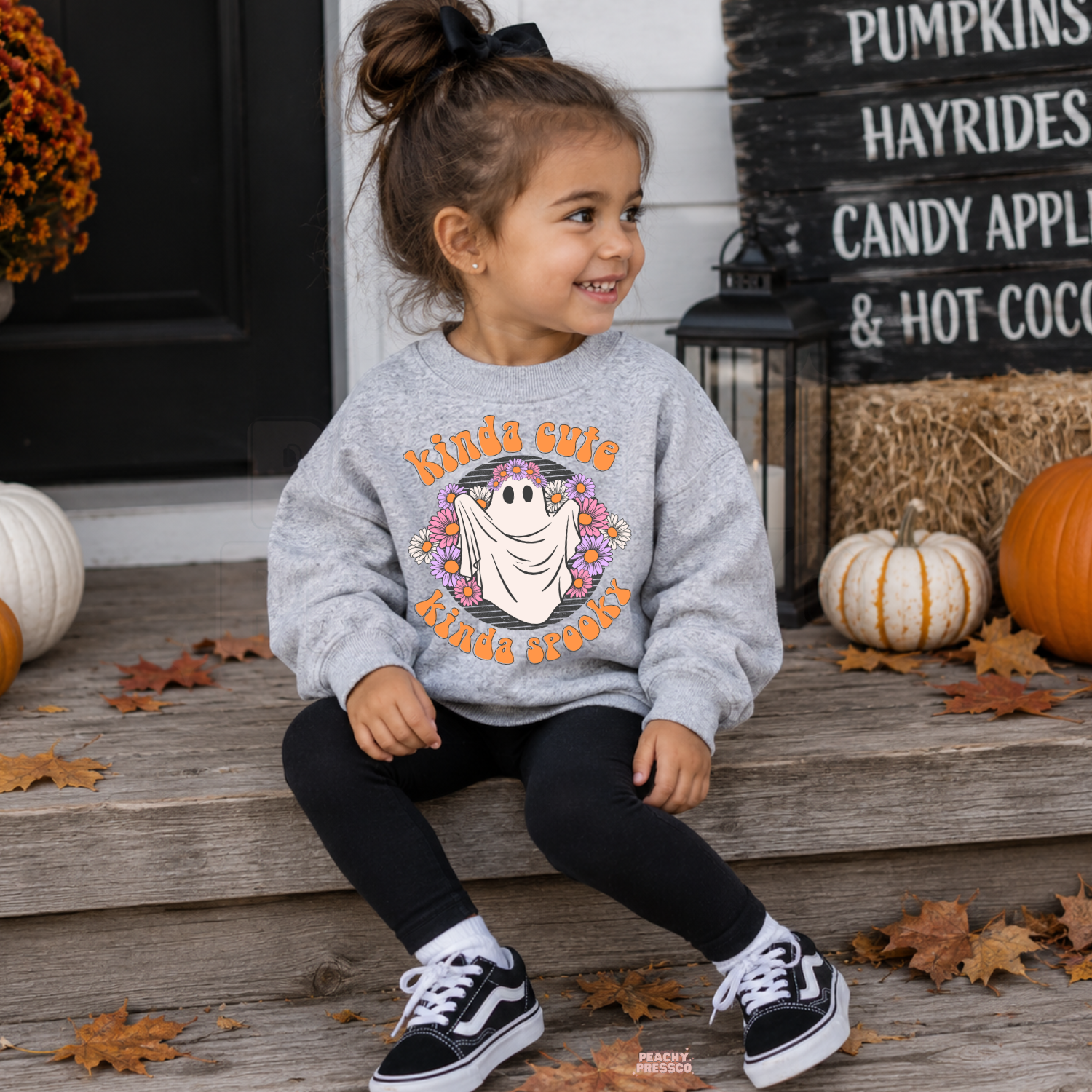Girl wearing a sport grey Kinda Cute Kinda Spooky Halloween sweatshirt with a floral ghost graphic and fall pumpkin decor.