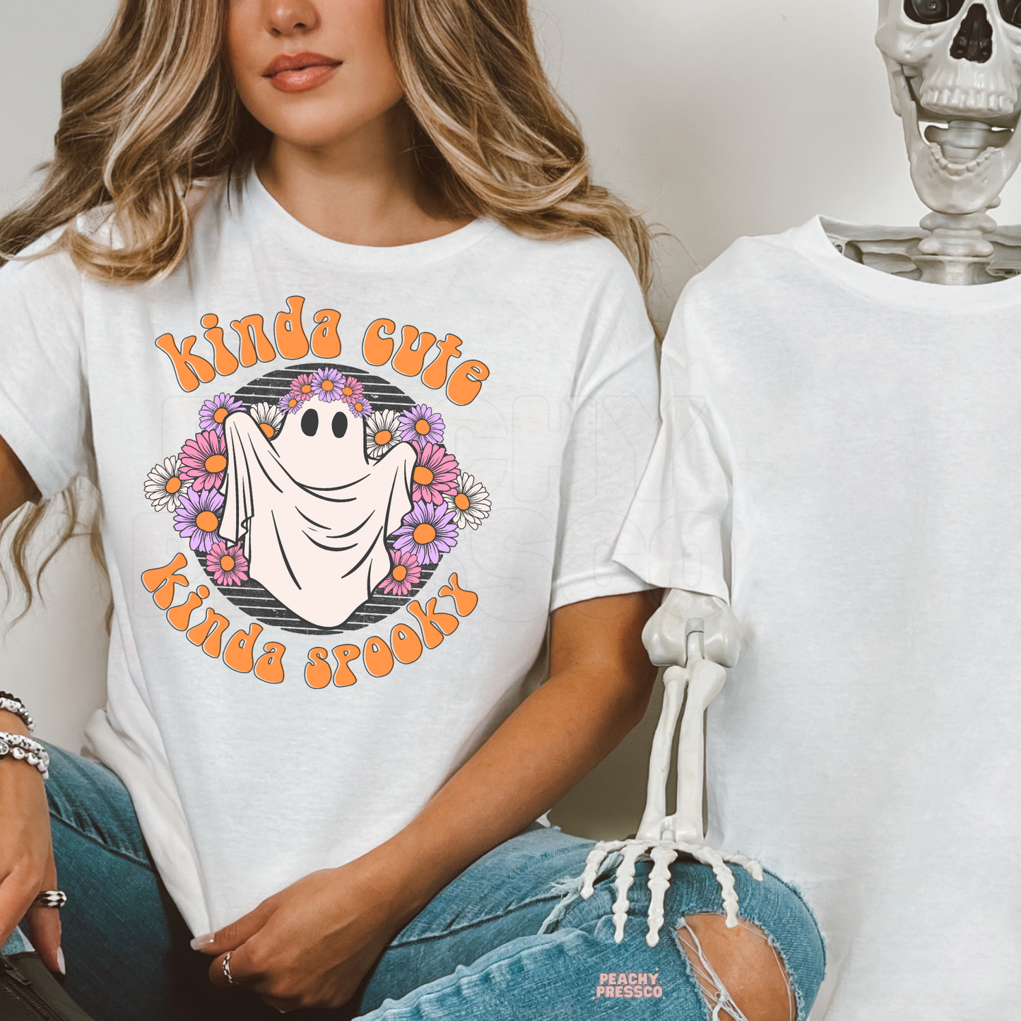 A woman wearing a white graphic t-shirt with a 'kinda cute kinda spooky' ghost design, posed next to a skeleton prop.
