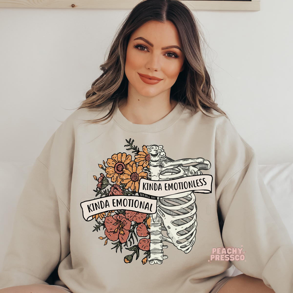 Sand crewneck sweatshirt with a floral skeleton ribcage graphic and the phrase "Kinda Emotional Kinda Emotionless," combining vintage botanical and gothic aesthetics.