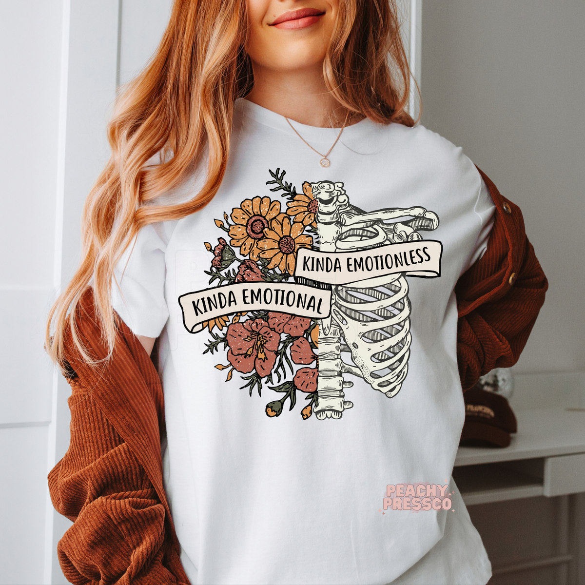 White t-shirt featuring a vintage anatomical ribcage with wildflowers and banner text reading "Kinda Emotional Kinda Emotionless," in a boho gothic floral style.
