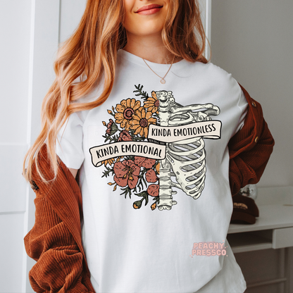 White t-shirt featuring a vintage anatomical ribcage with wildflowers and banner text reading "Kinda Emotional Kinda Emotionless," in a boho gothic floral style.