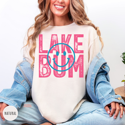 Natural graphic t-shirt with 'LAKE BUM' printed in distressed pink letters and a blue smiley face overlay, styled with a light blue denim jacket and jeans.