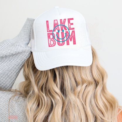 White mesh trucker hat featuring distressed pink “Lake Bum” lettering with turquoise smiley face design, perfect for lake days and summer adventures.