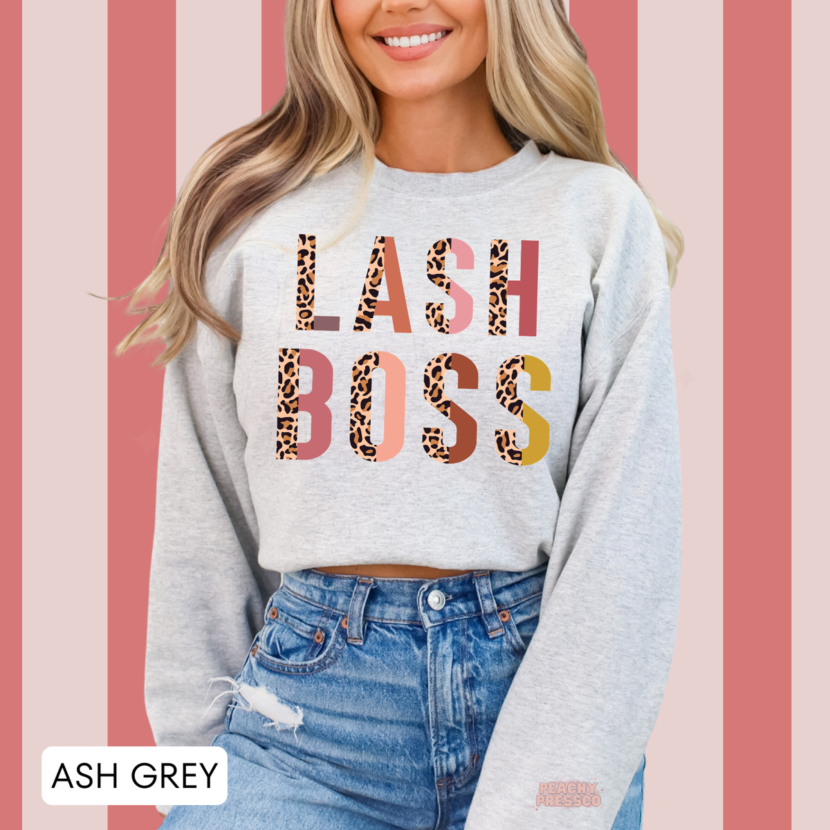 Woman modeling an ash grey Lash Boss crewneck sweatshirt featuring colorful leopard print lettering against a pink striped background.