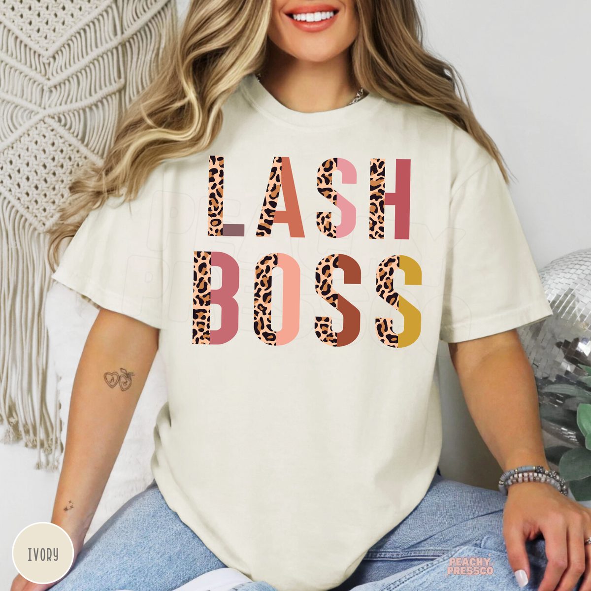 Woman modeling an ivory Lash Boss T-shirt featuring colorful leopard print lettering in a bold two-line design.