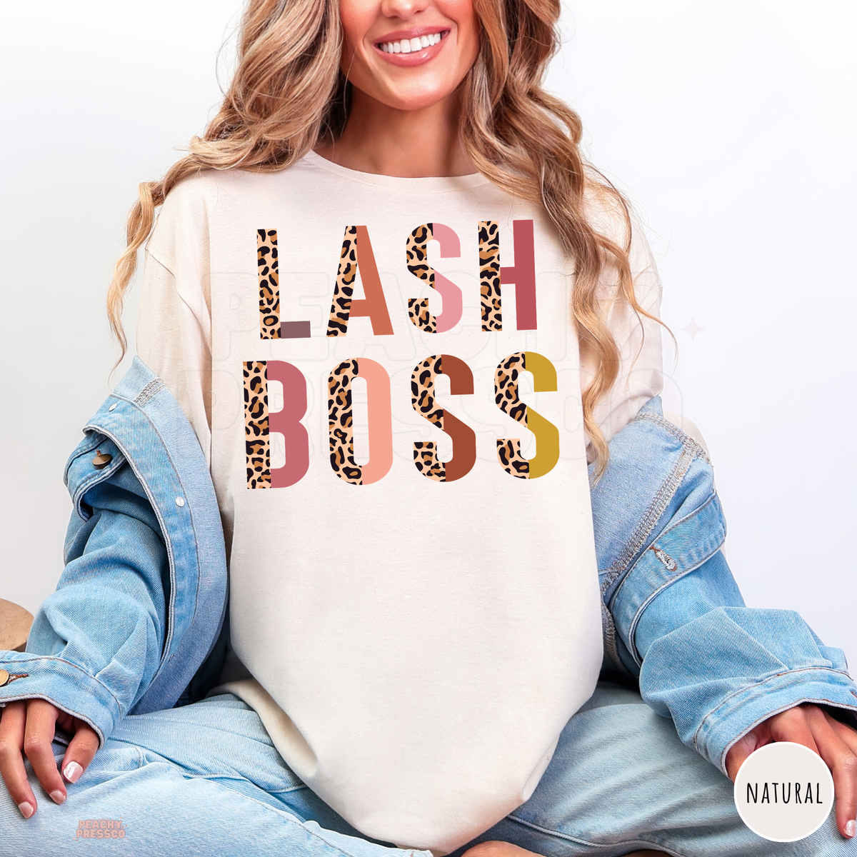 Woman wearing a natural colored Lash Boss graphic T-shirt featuring bold leopard print lettering with pink, coral and mustard accents.