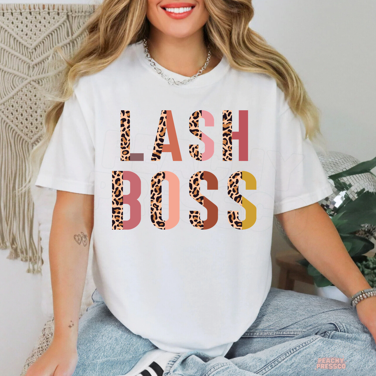 Woman wearing a white Lash Boss graphic T-shirt featuring trendy leopard print letters with pink, coral and mustard accents.
