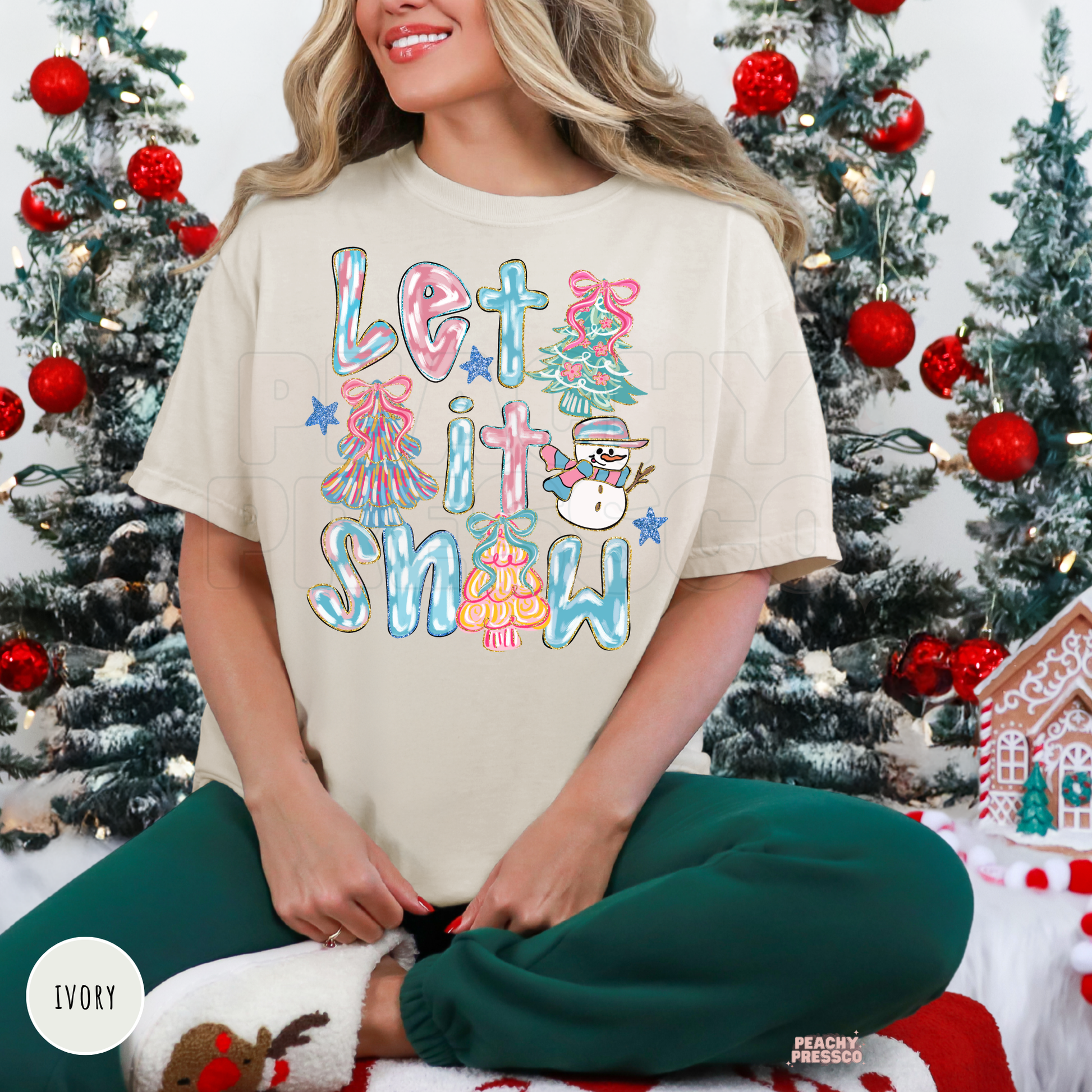 A woman sitting cross-legged in front of Christmas trees wearing an ivory graphic t-shirt that reads 'Let it Snow' in colorful pastel lettering with a snowman and tree illustration. She is wearing dark green pants and white slippers.