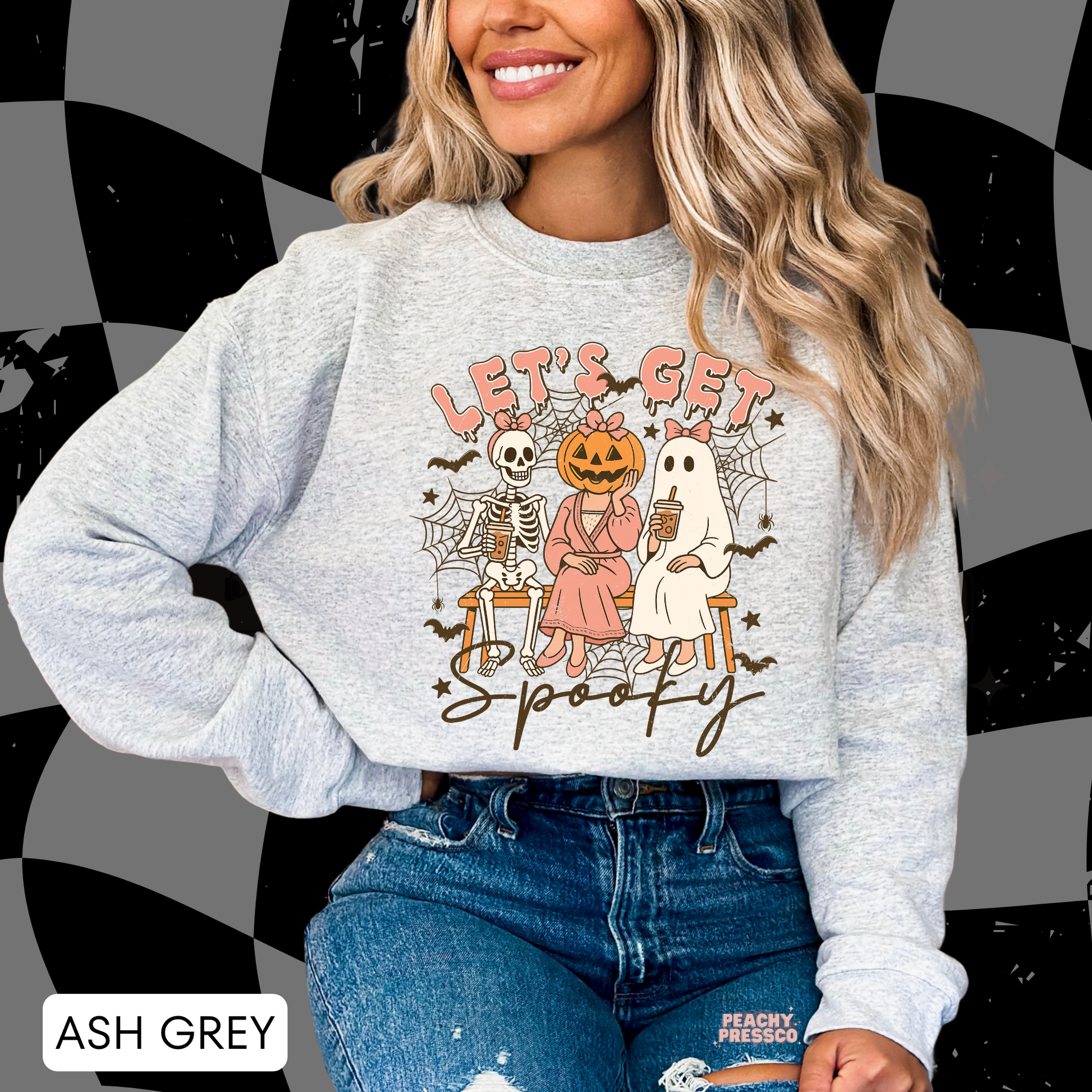 Woman wearing an ash grey crewneck sweatshirt featuring a cozy Let's Get Spooky Halloween graphic with a ghost, skeleton, pumpkin character and coffee.