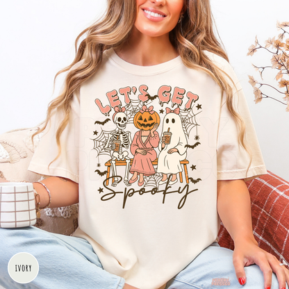 Woman wearing an ivory Comfort Colors t-shirt with a Let's Get Spooky graphic featuring a ghost, skeleton, pumpkin character, bats and spider webs.