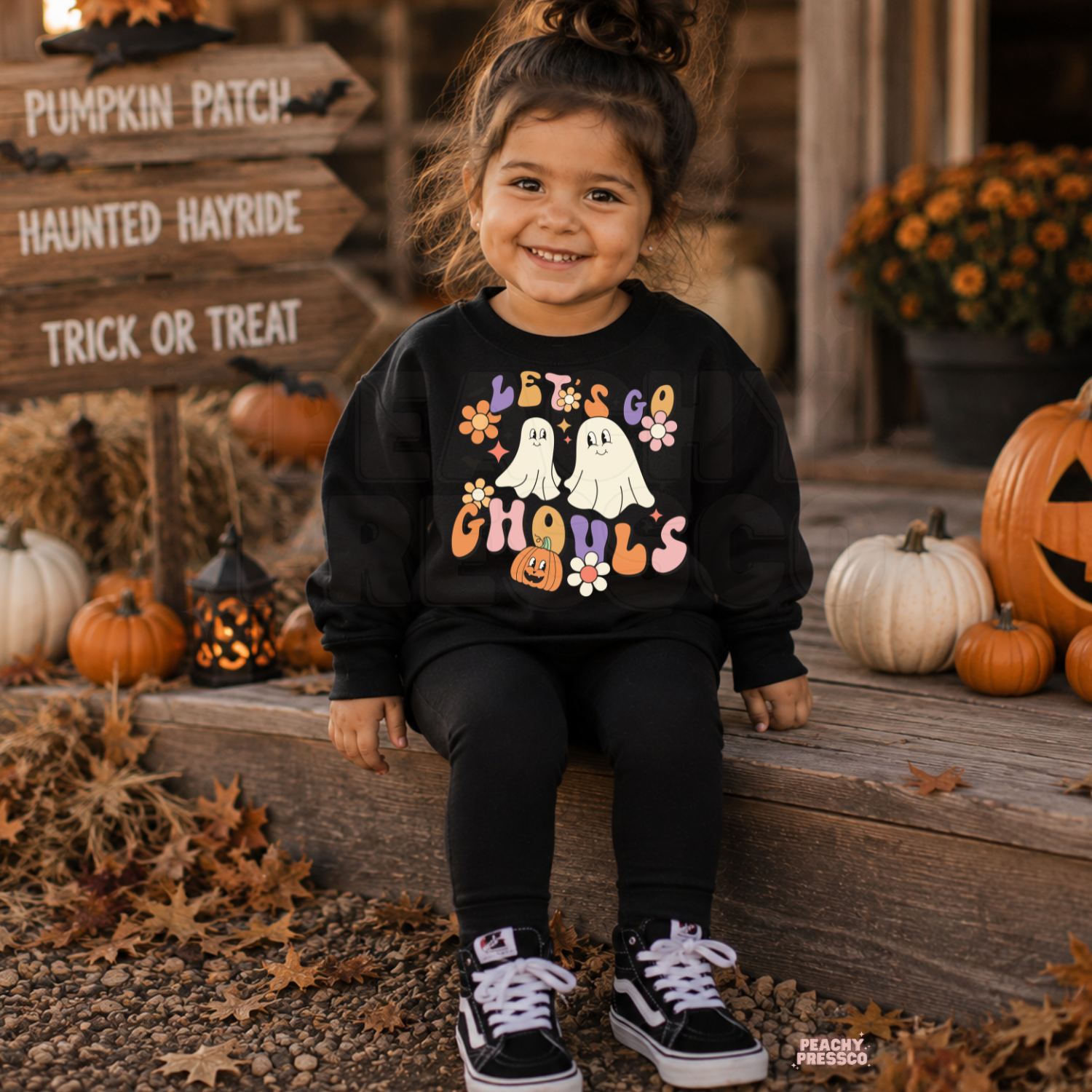 Girl wearing a black Let’s Go Ghouls Halloween sweatshirt with cute ghosts, colorful retro lettering, flowers and pumpkin design.