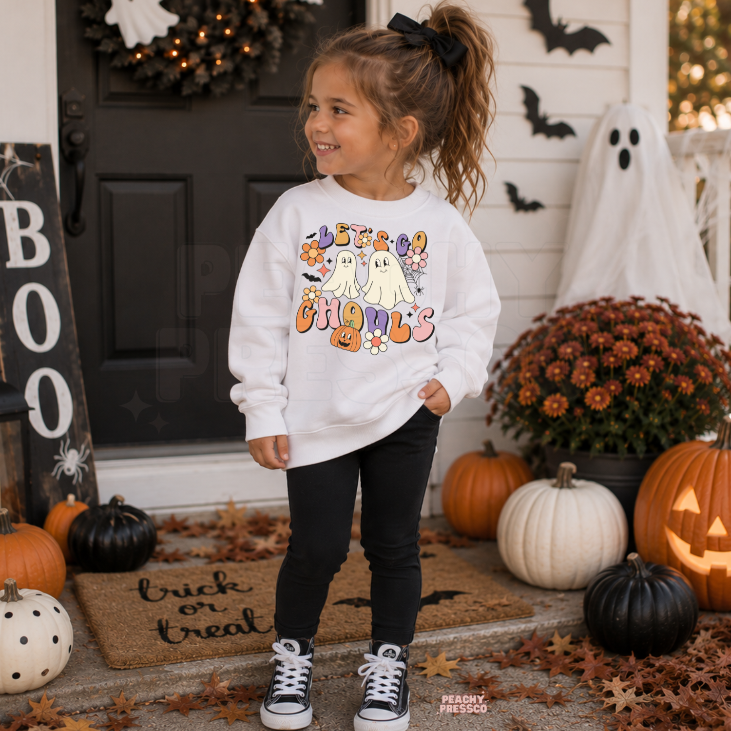 Girl wearing a white Let’s Go Ghouls Halloween sweatshirt featuring cute ghosts, colorful retro lettering, flowers and a pumpkin.
