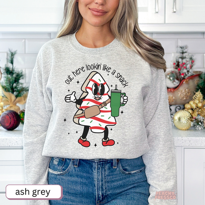 A woman wearing a ash grey sweatshirt with a cartoon candy cane graphic and the text 'out here lookin' like a snack'. The background shows a decorated kitchen with Christmas ornaments.