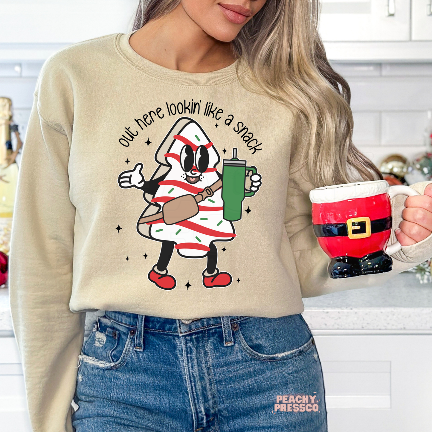 A woman wearing a beige crewneck sweatshirt with a cartoon character graphic that reads 'out here lookin' like a snack' and holding a red Santa-themed mug in a kitchen setting.