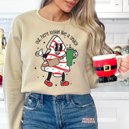 A woman wearing a beige crewneck sweatshirt with a cartoon character graphic that reads 'out here lookin' like a snack' and holding a red Santa-themed mug in a kitchen setting.