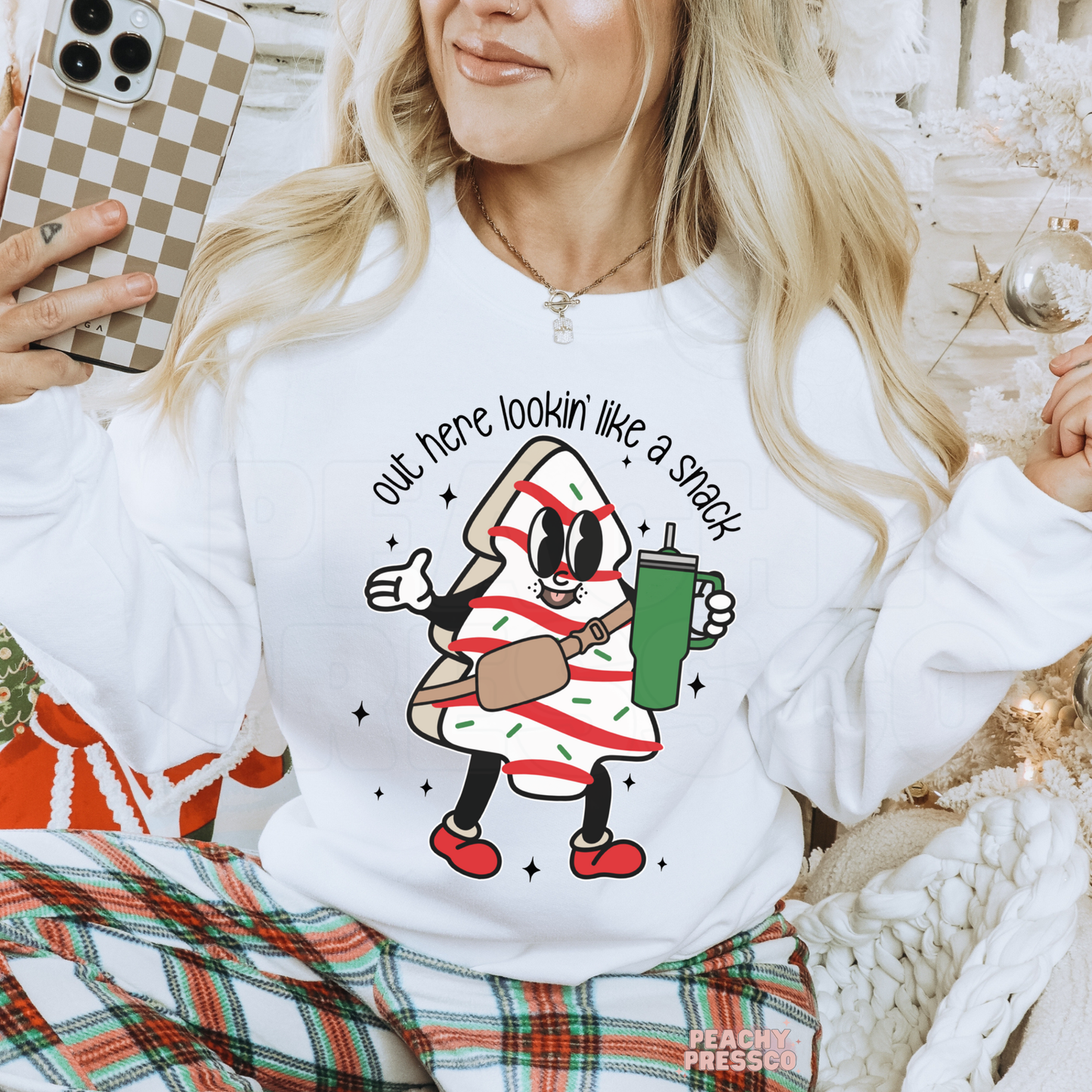 A woman wearing a white crewneck sweatshirt with a festive holiday graphic of a character dressed as a striped candy cane holding a green mug, with the text 'out here lookin' like a snack'. She is also wearing plaid pajama pants and holding a checkered phone case.