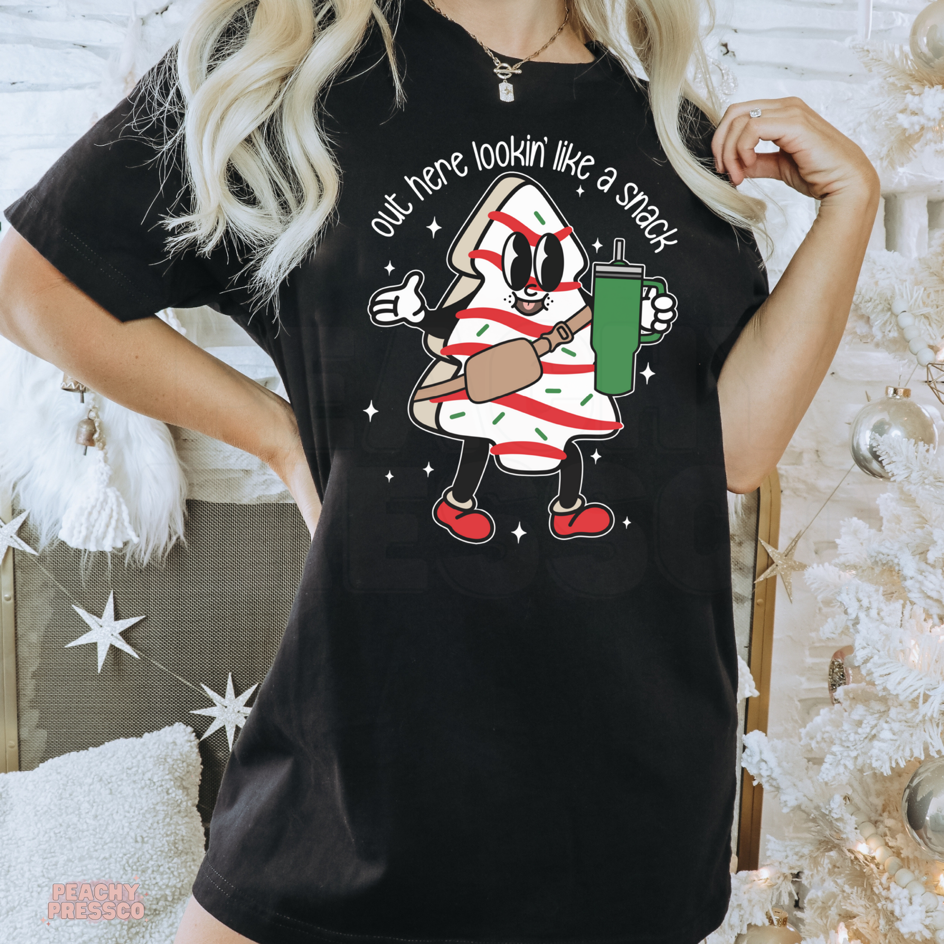 A woman wearing a black graphic t-shirt with a cartoon candy cane character and the text 'out here lookin' like a snack'. The background features a white Christmas tree and holiday decorations.