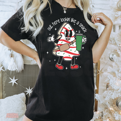 A woman wearing a black graphic t-shirt with a cartoon candy cane character and the text 'out here lookin' like a snack'. The background features a white Christmas tree and holiday decorations.
