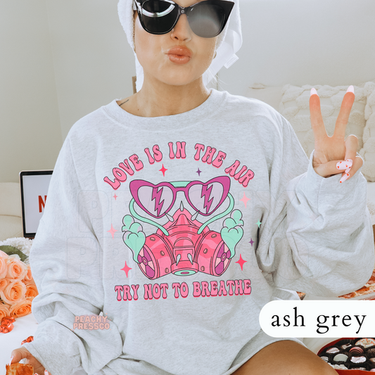 Love Is In The Air Try Not To Breathe design on an ash grey crewneck sweatshirt featuring a pink respirator mask, heart sunglasses, and retro Valentine's graphics.