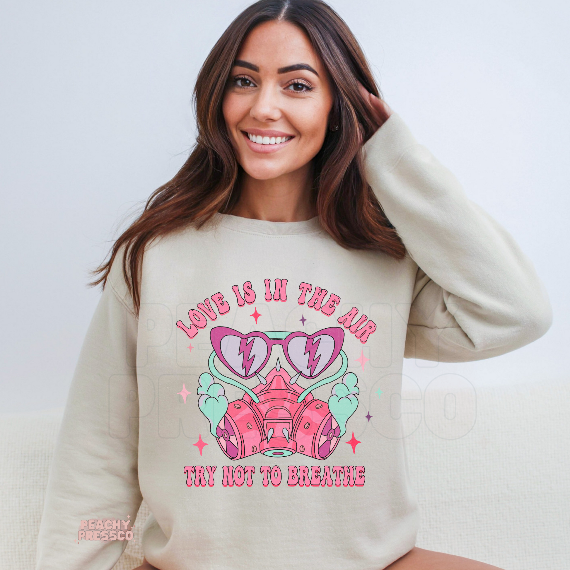 Love Is In The Air Try Not To Breathe graphic displayed on a sand-colored crewneck sweatshirt with retro respirator mask and playful Valentine's artwork.