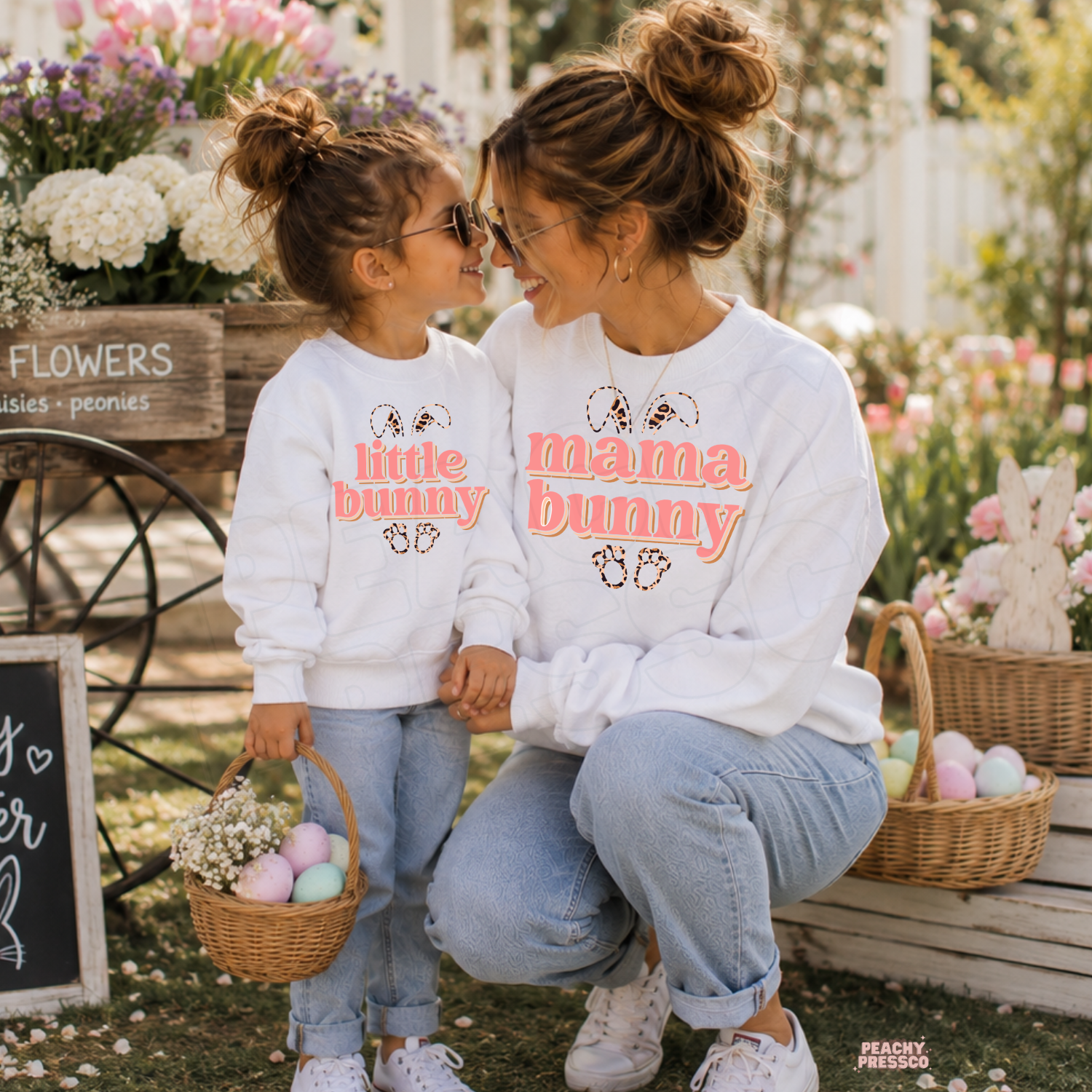 Mom and daughter wearing matching white Mama Bunny and Little Bunny Easter crewneck sweatshirts with pastel Easter baskets in a spring flower garden.
