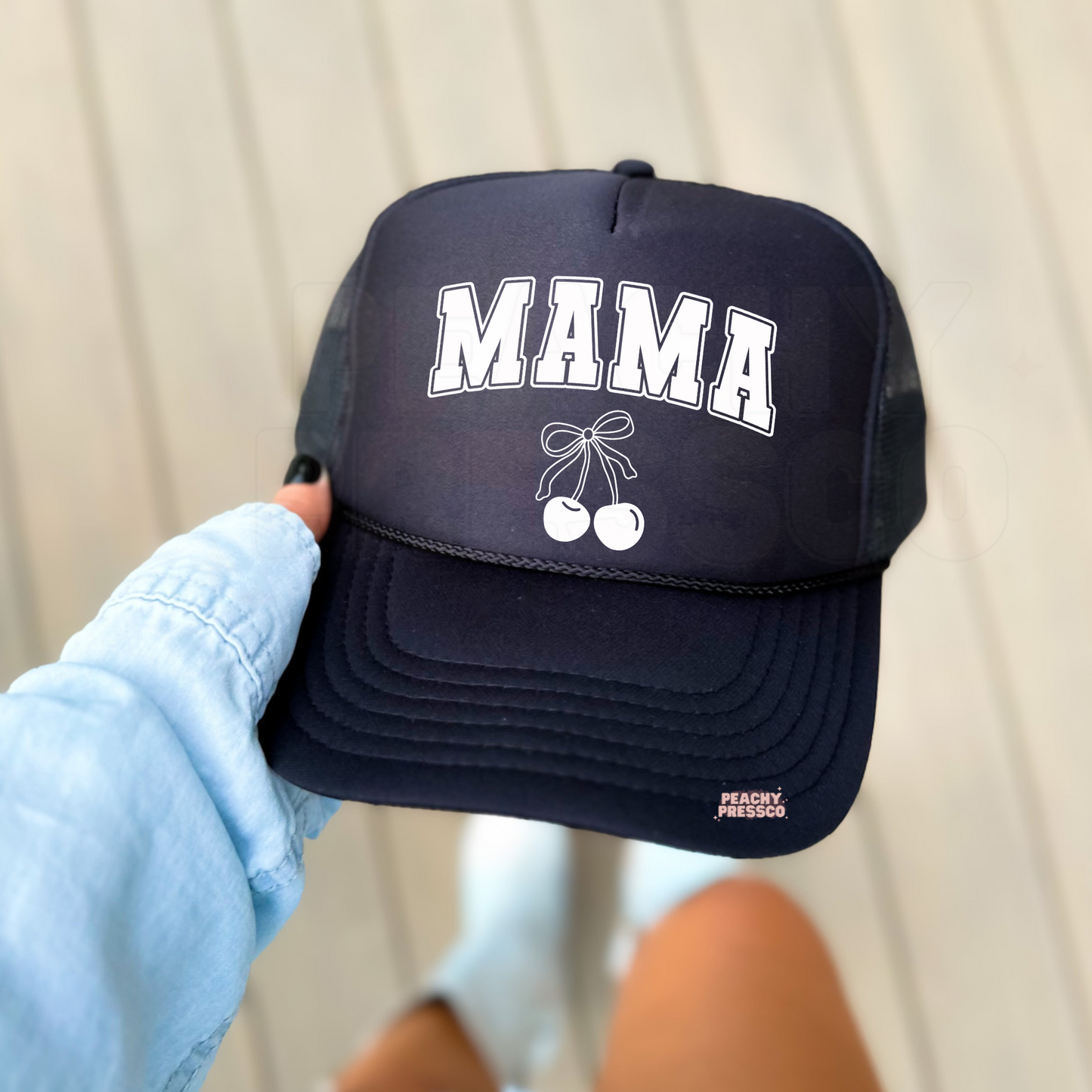 Black foam trucker hat featuring white varsity "MAMA" lettering and a white cherry bow graphic. Modern minimalist mom hat perfect for everyday wear, gifts for moms, errands, coffee runs, and casual outfits.