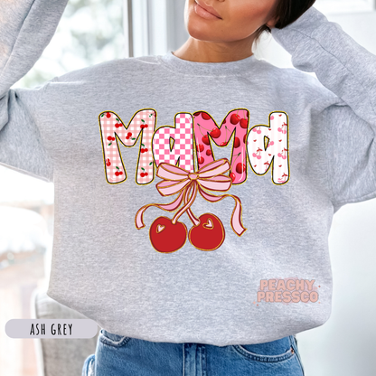 Woman wearing an ash grey sweatshirt featuring a Mama cherry bow graphic with pink checkerboard lettering and hanging cherries.