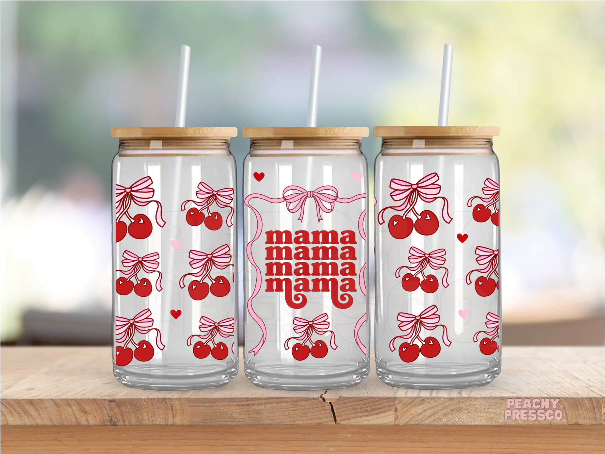 Glass cups with red cherry and bow patterns and the word 'mama' on them, each with a bamboo lid and a straw, displayed on a wooden surface with a blurred background.