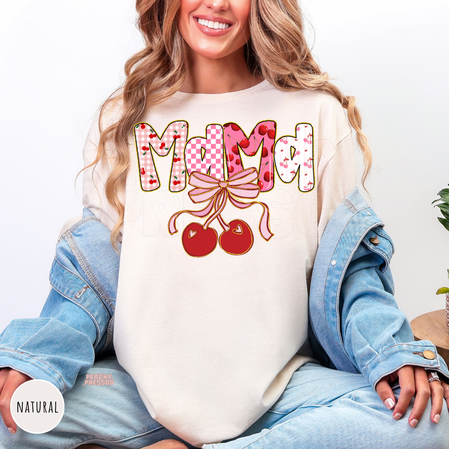 Woman wearing a natural shirt with a Mama cherry bow graphic featuring pink cherries, bows, and coquette-inspired lettering.