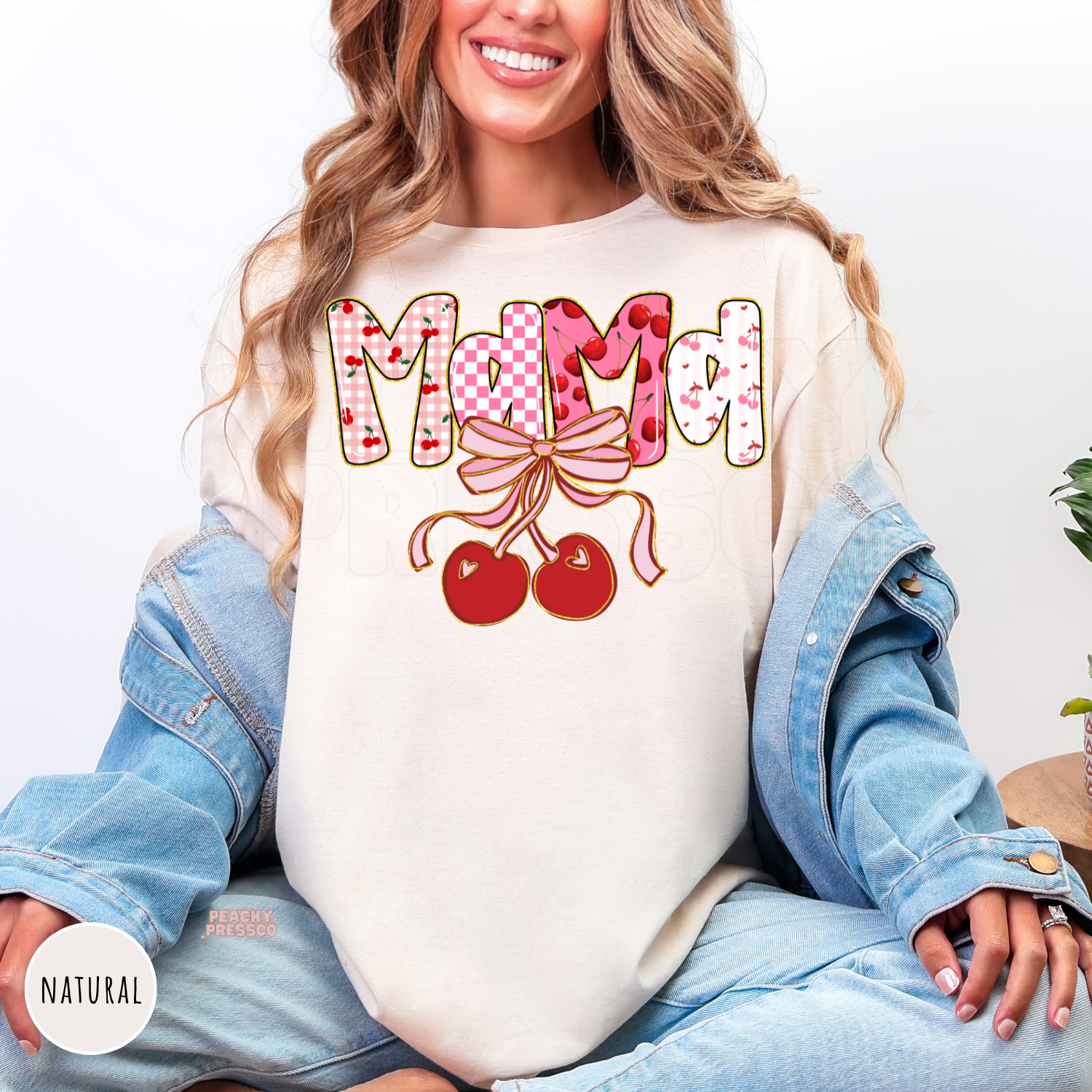 Woman wearing a natural shirt with a Mama cherry bow graphic featuring pink cherries, bows, and coquette-inspired lettering.