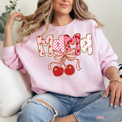 Woman wearing a light pink sweatshirt with a Mama cherry bow design featuring cherry print, checkerboard accents, and a pink ribbon.