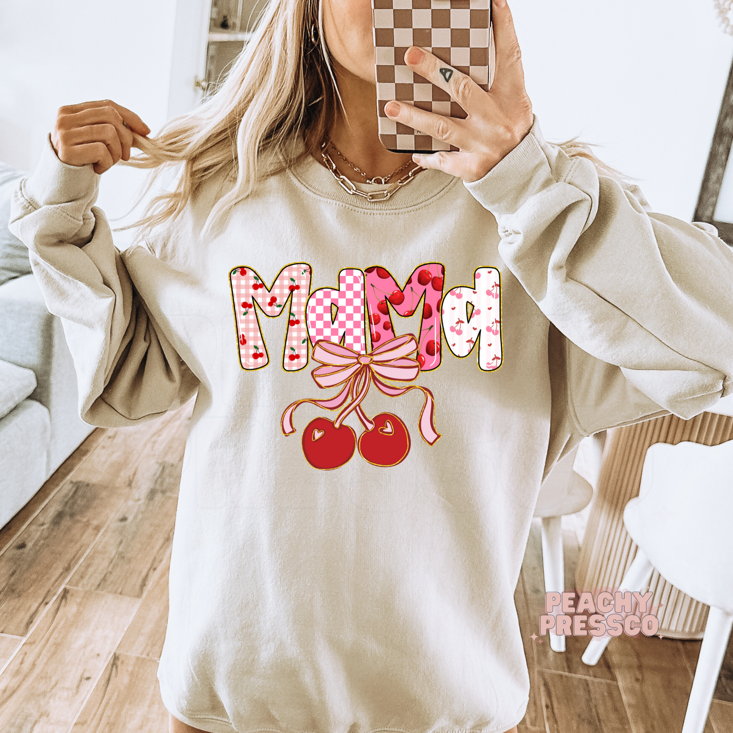 Mirror selfie of woman wearing a sand sweatshirt with a Mama cherry bow graphic in pink cherry print and checkerboard lettering.