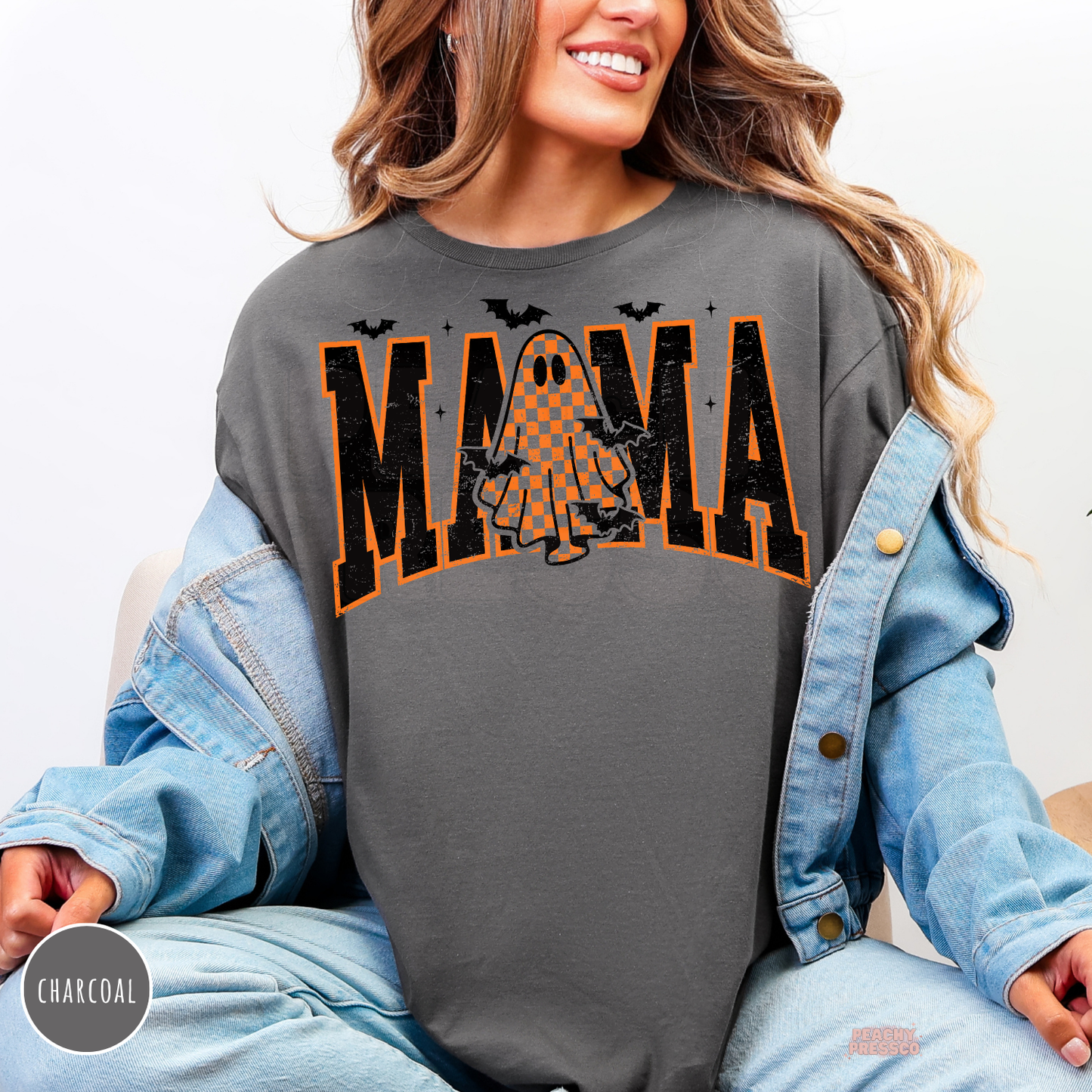 Woman wearing a charcoal crewneck sweatshirt with a distressed Mama Halloween graphic featuring an orange checkered ghost and spooky bats.