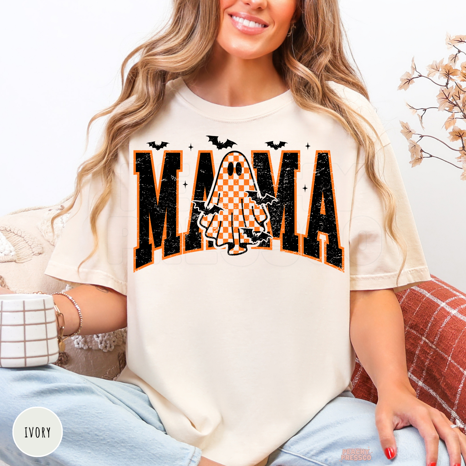Woman wearing an ivory Comfort Colors t-shirt with a distressed varsity Mama graphic featuring an orange checkered ghost and bats for Halloween.