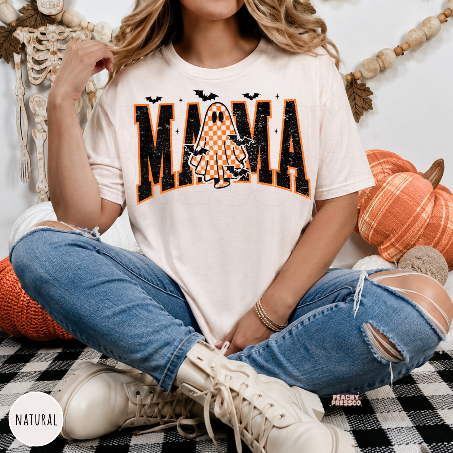Woman wearing a natural Comfort Colors t-shirt featuring a trendy Mama Halloween design with a checkered ghost, distressed varsity lettering and bats.