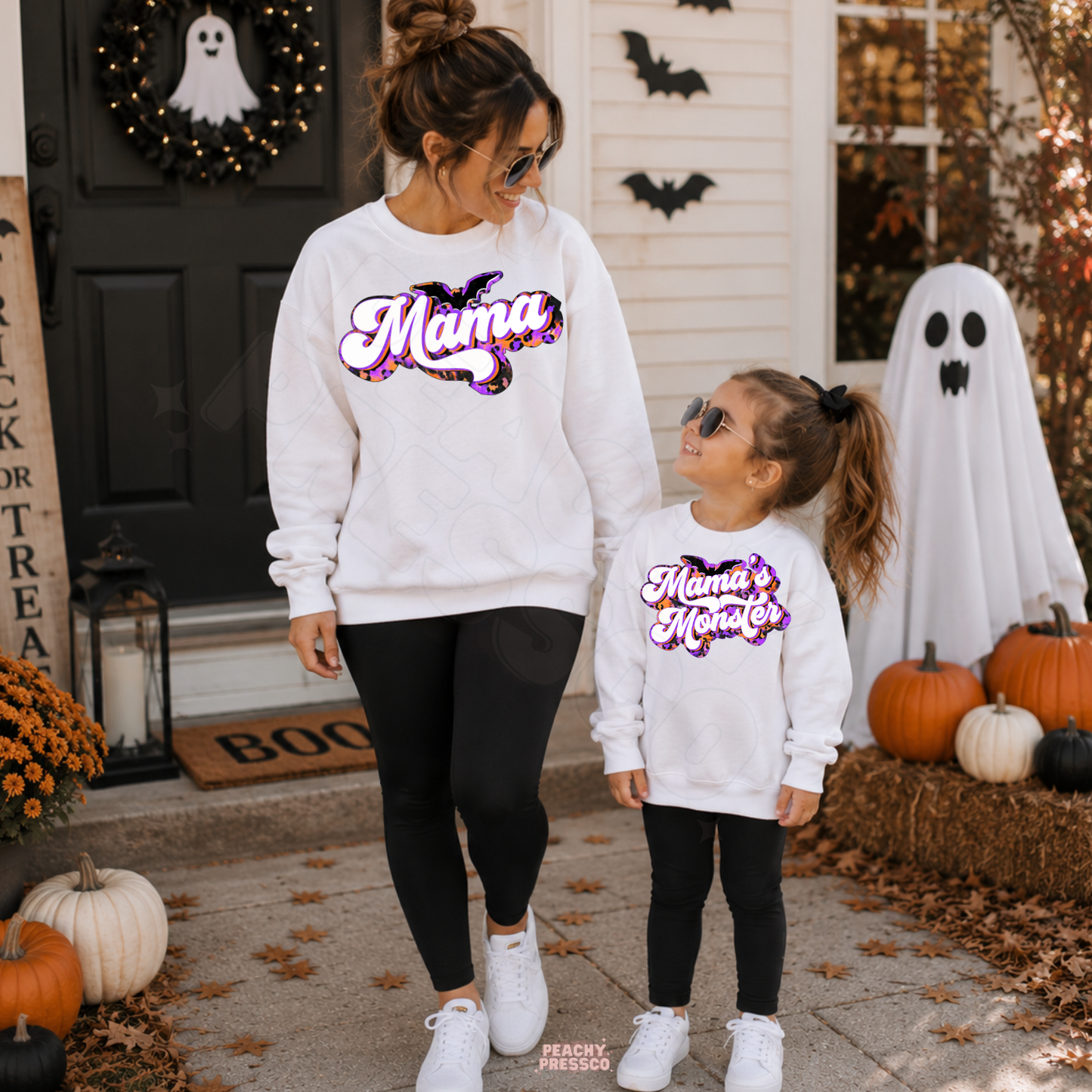 Mom and daughter wearing matching white Mama and Mama’s Monster Halloween sweatshirts surrounded by pumpkins, bats and ghost decor.