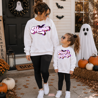 Mom and daughter wearing matching white Mama and Mama’s Monster Halloween sweatshirts surrounded by pumpkins, bats and ghost decor.